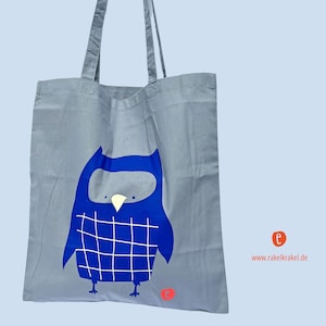 Könnte beinhalten: Graue Tragetasche mit einem blauen Eulenmuster. Die Eule hat einen weißen Schnabel und ein Gittermuster auf der Brust. Die Tasche hat lange graue Henkel und ein kleines orangefarbenes Logo. Die Website www.rakelkrakel.de ist sichtbar.