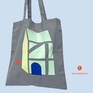 Könnte beinhalten: Eine graue Stofftasche mit einem Grafikdruck eines Hauses mit einer blauen Tür. Das Haus ist in Gelb und Grün umrandet. Der Text "www.rakelkrakel.de" ist auf der Tasche aufgedruckt.