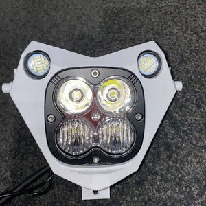 Puede incluir: Un conjunto de faros de motocicleta blancos con dos luces LED principales y dos luces auxiliares m&aacute;s peque&ntilde;as. El conjunto tiene un borde negro alrededor de las luces principales y un soporte de montaje.