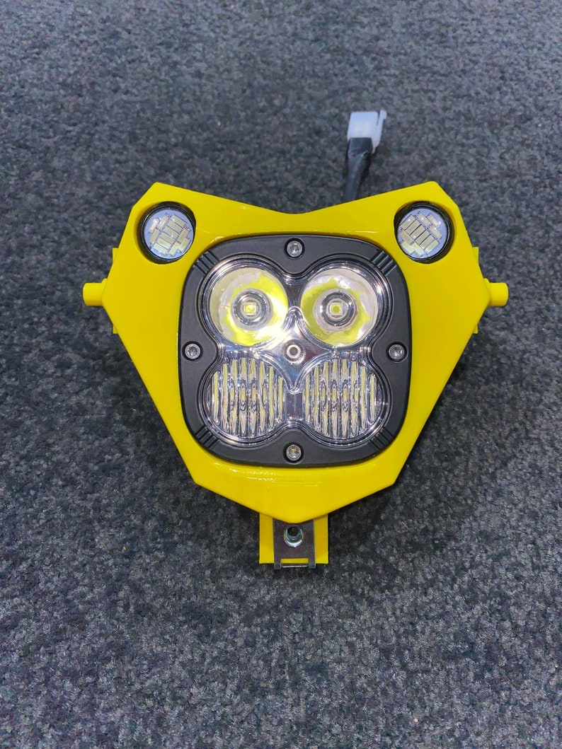Puede incluir: Un faro de motocicleta amarillo brillante con dos luces LED redondas en la parte superior y dos luces LED m&aacute;s grandes en la parte inferior. El conjunto tiene una secci&oacute;n central negra y un soporte de montaje en la parte inferior.