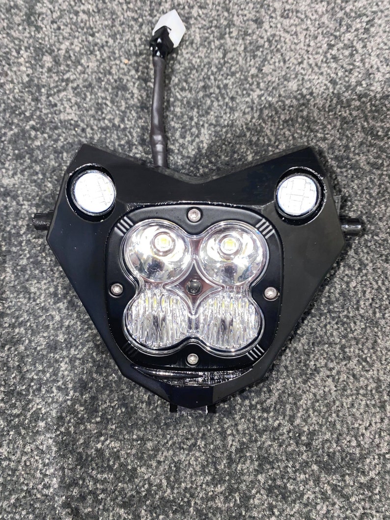 Puede incluir: Conjunto de faro de motocicleta negro con dos luces laterales redondas y cuatro luces LED centrales. El faro tiene una carcasa negra y un cable con un conector que se extiende desde la parte superior.