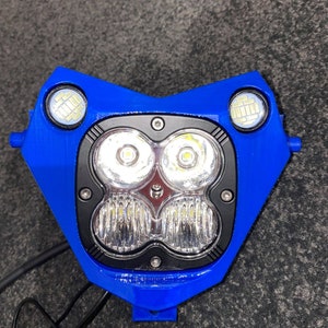 Puede incluir: Un conjunto de faros de motocicleta azules con m&uacute;ltiples luces LED. El conjunto tiene un dise&ntilde;o en forma de coraz&oacute;n con dos luces m&aacute;s peque&ntilde;as en la parte superior y un grupo de luces m&aacute;s grandes en el centro. El marco es azul y las luces son de color blanco brillante.