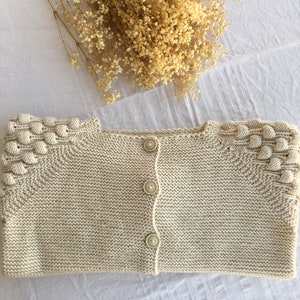 Pode incluir: Um cardigan de malha creme com fecho de botões e um design texturizado nos ombros.