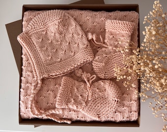 May include: A baby gift set in a brown box, featuring a knitted blanket, bonnet, and booties in a soft, light pink color. The set has a textured pattern and is ideal for a newborn.