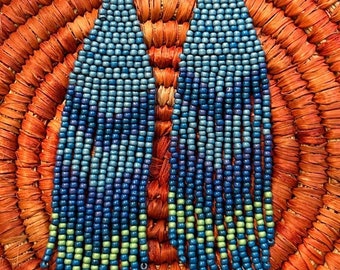 Könnte beinhalten: Ein Paar lange, geperlte Ohrringe mit einem Farbverlauf aus blauen, grünen und violetten Perlen. Die Ohrringe hängen an silbernen Haken und werden auf einem gewebten orangefarbenen Hintergrund präsentiert. Der Preis beträgt 40 €.