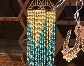 Könnte beinhalten: Ein Paar lange, geperlte Ohrringe mit einem goldenen Sonnenstrahl-Design an der Oberseite. Die Perlen sind in einer Mischung aus gelben und blauen Farben.