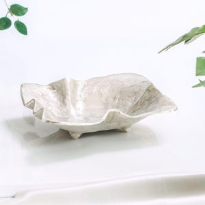 May include: A white ceramic bowl with a unique, organic shape and a textured surface. The bowl has four small feet and is perfect for serving snacks or holding small items.