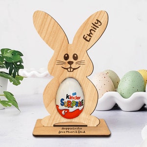 Op de afbeelding: Houten paashazen-eierhouder met de gepersonaliseerde naam "Emily" en de tekst "Prettige Pasen Liefs Mama & Papa". De haas houdt een Kinder Surprise-ei vast.