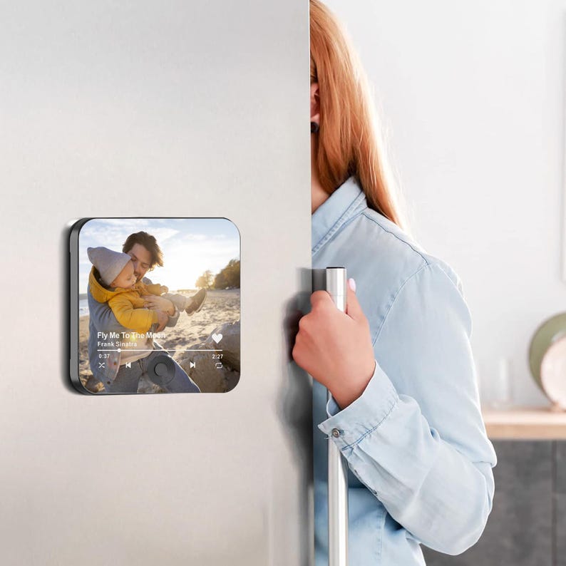 May include: A square magnet with rounded corners, featuring a photo of a father and child on a beach. The magnet has a black border and displays the song title "Fly Me To The Moon" by Frank Sinatra.