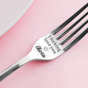 May include: A silver fork engraved with the words "I forking love you" and the name "Olivia". A heart and sun design is also engraved. The fork is set against a pink backdrop.