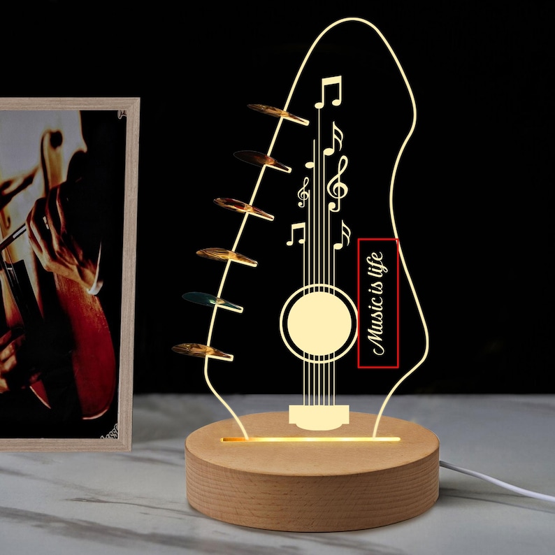 Pu&ograve; includere: Lampada a LED a forma di chitarra con note musicali e la scritta "Music is life". La lampada ha una base in legno e una forma di chitarra in acrilico trasparente con una calda luce gialla. Sullo sfondo c'&egrave; una foto incorniciata di un musicista.
