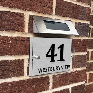 May include: A silver house number sign with black lettering. The sign reads "41 WESTBURY VIEW". The sign is mounted on a brick wall with a solar light above it.