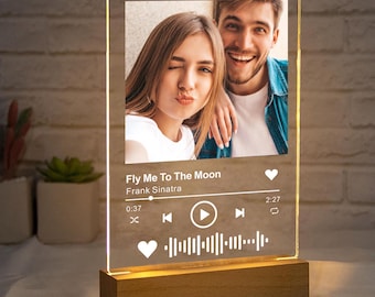 May include: A clear acrylic photo frame with a couple's photo and a music player design. The music player has a song title "Fly Me To The Moon" by Frank Sinatra, a play button, and a progress bar. The frame is on a wooden base.