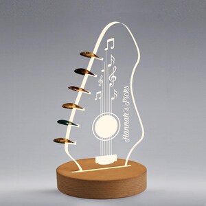 Pu&ograve; includere: Lampada a LED a forma di chitarra con base in legno. La lampada presenta un contorno bianco di una chitarra con note musicali e la scritta "Hannah's Picks". Sei plettri sono attaccati al lato. La lampada &egrave; illuminata da una calda luce.