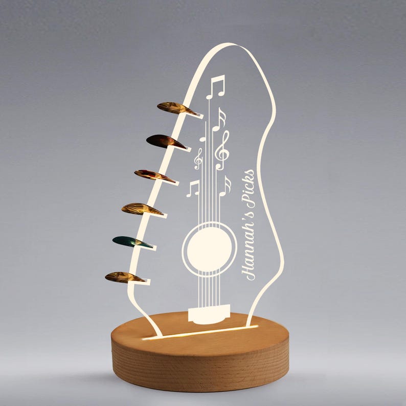 Pu&ograve; includere: Lampada a LED a forma di chitarra con base in legno. La lampada presenta un contorno bianco di una chitarra con note musicali e la scritta "Hannah's Picks". Sei plettri sono attaccati al lato. La lampada &egrave; illuminata da una calda luce.