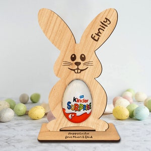 Op de afbeelding: Een houten paas konijnenstandaard met een gepersonaliseerde naam "Emily" en de tekst "Prettige Pasen Liefs Mama & Papa". De standaard houdt een Kinder Surprise chocolade-ei.