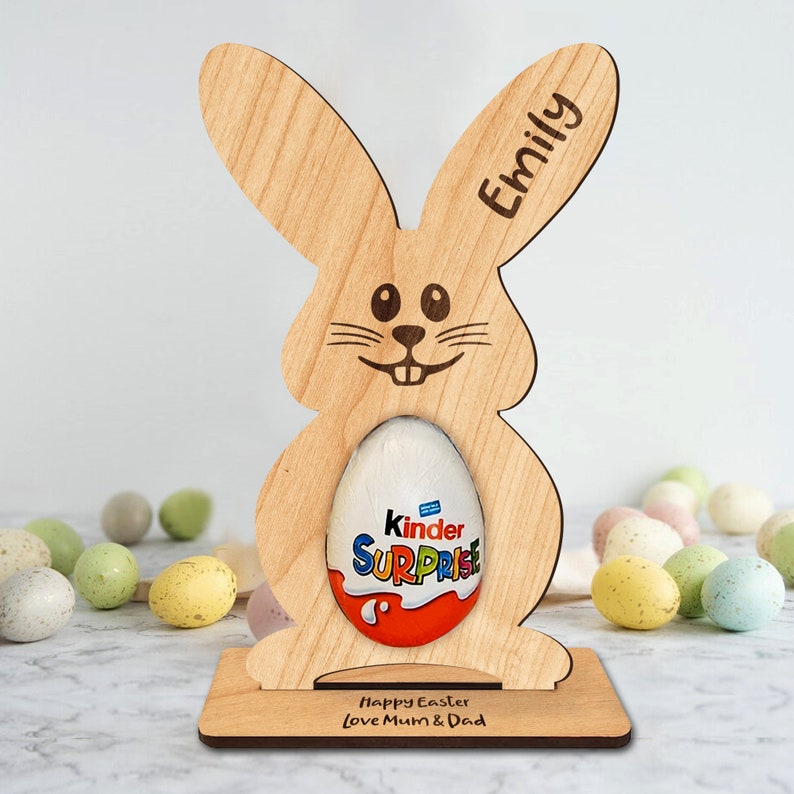 Op de afbeelding: Een houten paas konijnenstandaard met een gepersonaliseerde naam "Emily" en de tekst "Prettige Pasen Liefs Mama & Papa". De standaard houdt een Kinder Surprise chocolade-ei.
