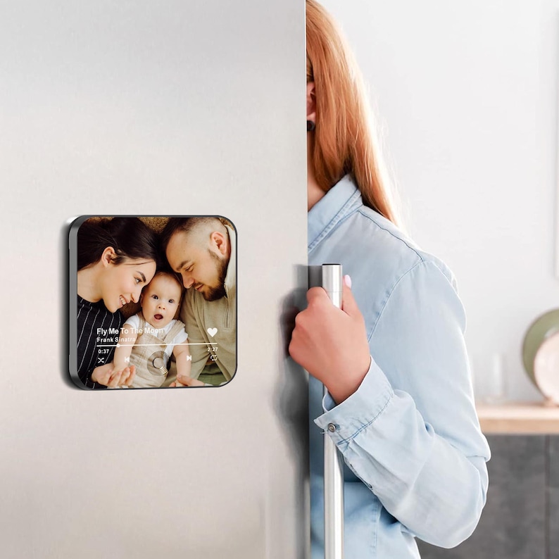 May include: A square magnet with a family photo and music player interface is displayed on a refrigerator. The photo shows a smiling family with a baby. The music player shows the song "Fly Me To The Moon" by Frank Sinatra.