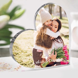 May include: A heart-shaped clear acrylic photo frame with a picture of a mother and daughter hugging. The photo is taken outdoors and the daughter is wearing a white hat and a brown fur vest. The mother is holding a bouquet of pink tulips. The text "Lydia & Danny" is engraved on the bottom of the frame.