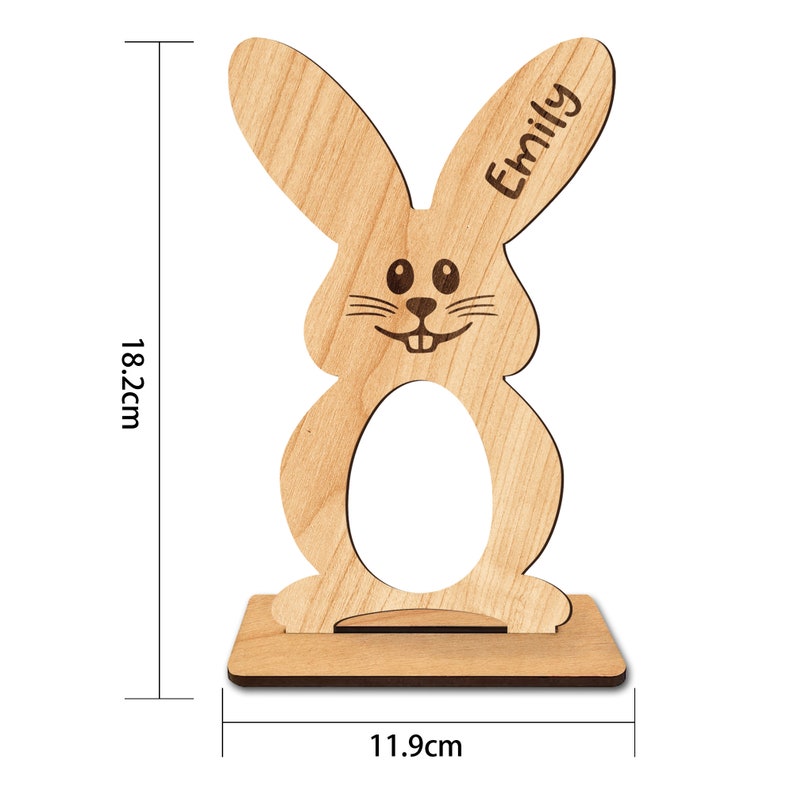 Op de afbeelding: Houten paaseierenhouder in de vorm van een konijn met een uitsparing voor een ei. Het konijn heeft een glimlachend gezicht en de naam "Emily" is in het oor gegraveerd. De houder is 18,2 cm hoog en 11,9 cm breed.