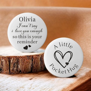 May include: Two white pocket hug tokens with black text. One says "Olivia I can't say I love you enough so this is your reminder" with two hearts. The other says "A little Pocket Hug" with a heart.