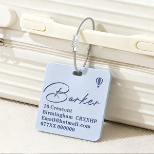 May include: A light blue square luggage tag with a metal cable loop. The tag features a dark blue hot air balloon icon and the text: Barker, 10 Crescent, Birmingham CRXXHP, Email@hotmail.com, 077XX 000000. The tag is attached to a cream-coloured suitcase handle.