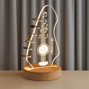 Pu&ograve; includere: Lampada a LED a forma di chitarra con base in legno. La lampada presenta note musicali e la scritta "Music is life". La paletta della chitarra ha pioli decorativi. La lampada emette una calda luce, ideale per gli amanti della musica.