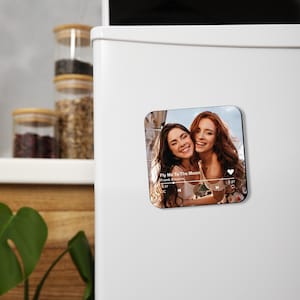 May include: Square magnet featuring a photo of two smiling women. The magnet includes a music player interface with the song title "Fly Me To The Moon" by Frank Sinatra. The background is a blurred outdoor scene.