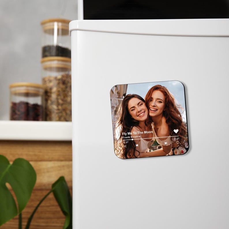 May include: Square magnet featuring a photo of two smiling women. The magnet includes a music player interface with the song title "Fly Me To The Moon" by Frank Sinatra. The background is a blurred outdoor scene.