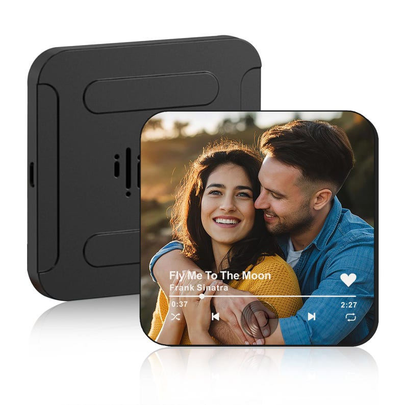 May include: Black square device with a screen displaying a photo of a smiling couple. The screen shows a music player interface with the song "Fly Me To The Moon" by Frank Sinatra. The device has a speaker and a stand.