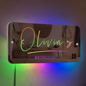 May include: A personalised bedroom sign with the name "Olivia's" written in rainbow colours. The sign is illuminated with colourful LED lights and has the word "BEDROOM" printed below the name.