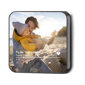May include: A black square digital photo frame displays a photo of a father holding a baby on a beach. The frame shows a music player interface with the song "Fly Me To The Moon" by Frank Sinatra.