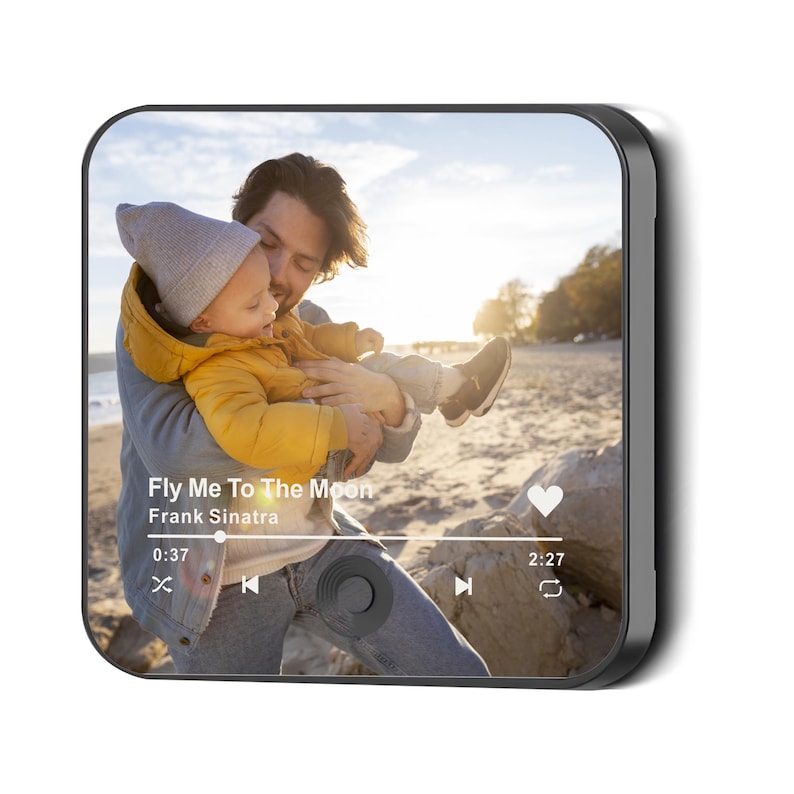 May include: A black square digital photo frame displays a photo of a father holding a baby on a beach. The frame shows a music player interface with the song "Fly Me To The Moon" by Frank Sinatra.