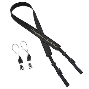 May include: Black leather camera strap with gold lettering that reads "Through the lens, See the World". The strap has two black adjustable clips.