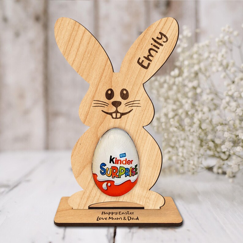 Op de afbeelding: Houten paashaas eierenhouder met de naam "Emily" gegraveerd op het oor. De houder heeft een gleuf voor een Kinder Surprise ei en een basis met de tekst "Prettige Pasen Liefs Mama & Papa".