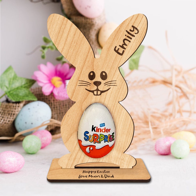 Op de afbeelding: Houten paashazen-eierhouder met een Kinder Surprise chocolade-ei erin. De houder heeft de naam "Emily" gegraveerd op het oor en de tekst "Prettige Pasen Liefs Mama & Papa" op de basis.