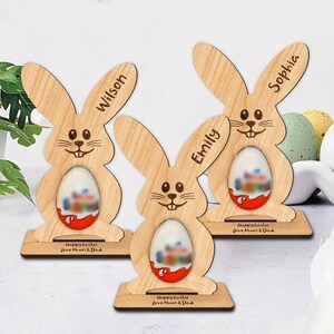 Op de afbeelding: Drie houten paashazen-eierhouders met de gepersonaliseerde namen "Wilson", "Emily" en "Sophia". Elke haas heeft een gleuf voor een chocolade-ei en een voet met de tekst "Prettige Pasen Liefs Mama en Papa".
