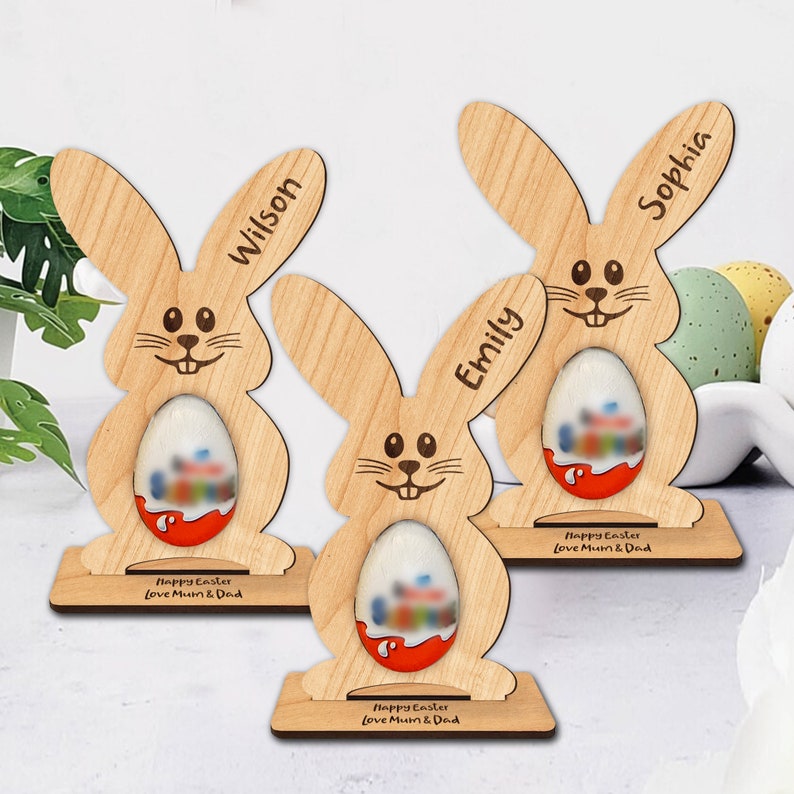 Op de afbeelding: Drie houten paashazen-eierhouders met de gepersonaliseerde namen "Wilson", "Emily" en "Sophia". Elke haas heeft een gleuf voor een chocolade-ei en een voet met de tekst "Prettige Pasen Liefs Mama en Papa".