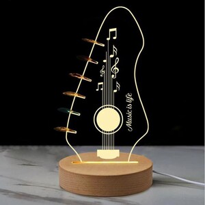 Pu&ograve; includere: Lampada a LED a forma di chitarra con base in legno. La lampada presenta un design a pentagramma musicale con note e la scritta "Music is life". La lampada &egrave; illuminata da una luce calda e soffusa, creando un'atmosfera decorativa e artistica.