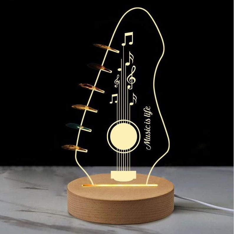 Pu&ograve; includere: Lampada a LED a forma di chitarra con base in legno. La lampada presenta un design a pentagramma musicale con note e la scritta "Music is life". La lampada &egrave; illuminata da una luce calda e soffusa, creando un'atmosfera decorativa e artistica.
