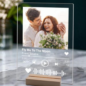 May include: A clear acrylic photo frame featuring a couple's photo and a music player interface. The music player displays the song "Fly Me To The Moon" by Frank Sinatra. The frame has a wooden stand.