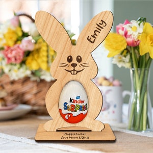 Op de afbeelding: Houten paashazen-eierhouder met een Kinder Surprise-ei erin. De haas heeft een glimlachend gezicht en de woorden "Emily" en "Prettige Pasen Liefs Mama & Papa" zijn gegraveerd op de basis.