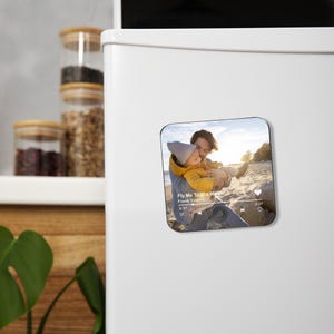 May include: Square magnet featuring a photo of a person holding a child on a beach. The magnet has a music player interface with the text "Fly Me To The Moon" by Frank Sinatra. The background shows a sunny beach scene.