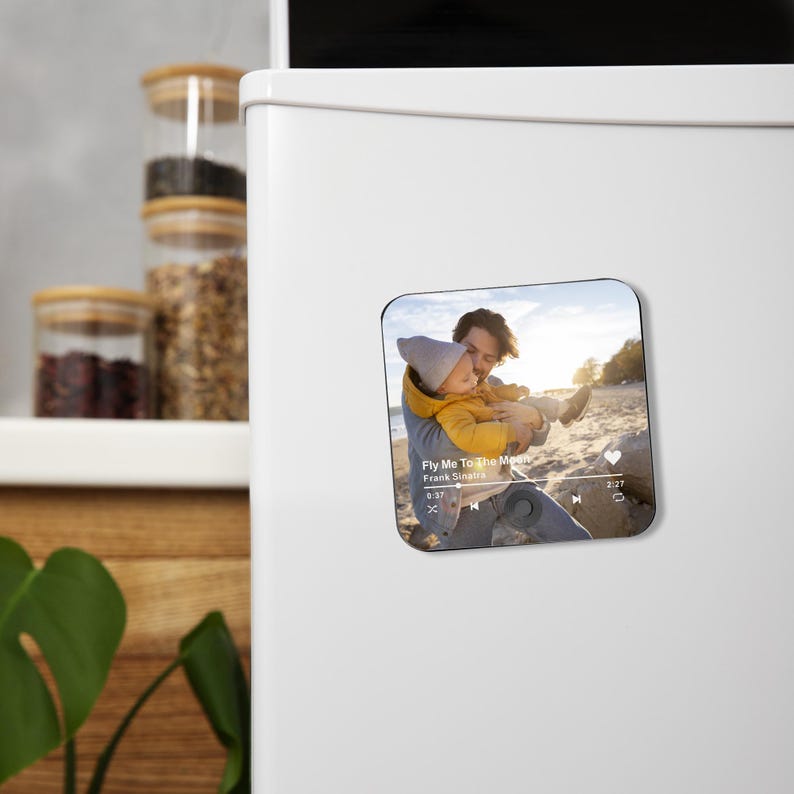 May include: Square magnet featuring a photo of a person holding a child on a beach. The magnet has a music player interface with the text "Fly Me To The Moon" by Frank Sinatra. The background shows a sunny beach scene.