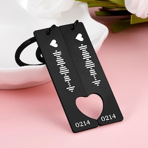 May include: Two black keyrings with a heart-shaped cutout and the number "0214". Each keyring has a white heart and a white sound wave pattern. The keyrings are connected by a black key ring.