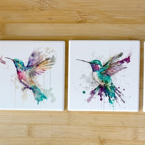 May include: Set of four ceramic coasters featuring watercolor paintings of hummingbirds with splashes of color. Each coaster depicts a hummingbird in a different pose with vibrant green, blue, and purple hues.