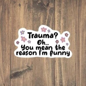May include: White sticker with a black outline and pink flowers. The text on the sticker reads "Trauma? Oh... You mean the reason I'm funny."