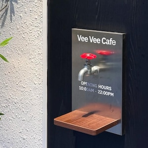 Puede incluir: Un letrero para Vee Vee Cafe con un grifo y un mango rojo. El letrero incluye el nombre del café y el horario de apertura, de 10:00 a 22:00. Una pequeña repisa de madera está debajo del letrero.