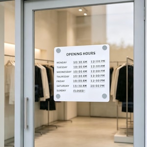 May include: A white rectangular sign with rounded corners attached to a glass door, displaying the text OPENING HOURS followed by a list of days from Monday to Sunday with specific times, including 10:30 AM to 12:00 PM and closed on Sunday.