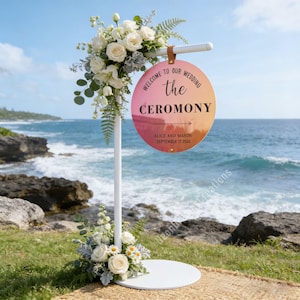 Puede incluir: Un letrero blanco para la ceremonia de boda con decoraciones florales. El letrero redondo tiene un fondo degradado rosa y naranja y dice "Welcome to our wedding the ceremony" con los nombres Alice y Mason y la fecha 17 de septiembre de 2026.